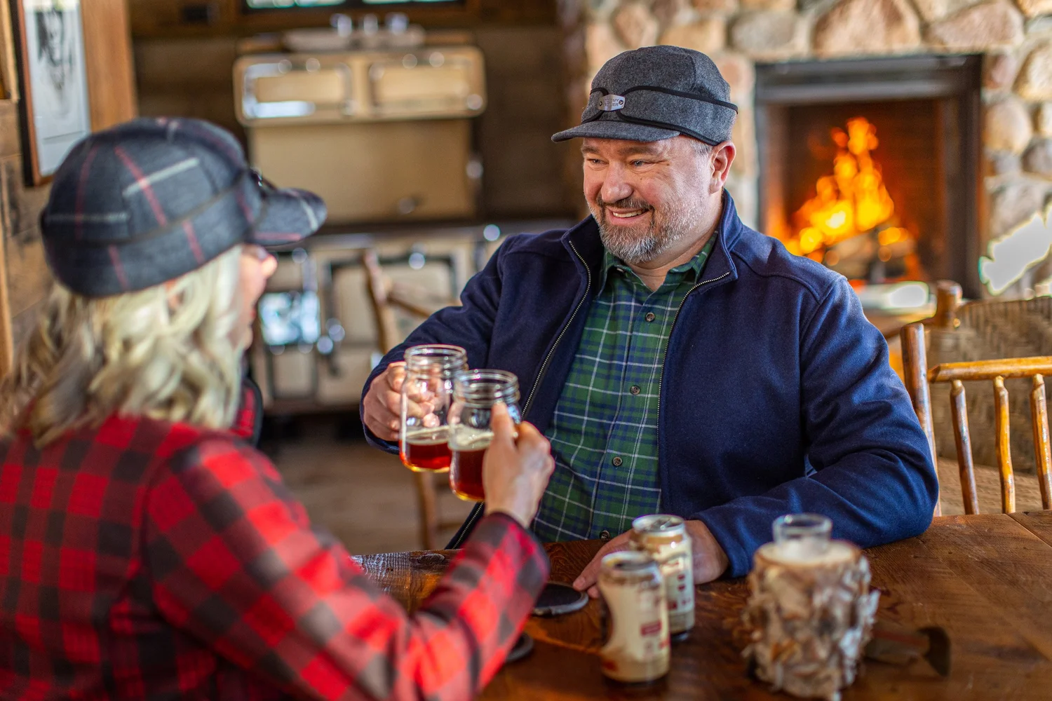 Two people in plaid caps raise glass mugs of beer in a toast at a cabin table, fireplace glowing behind